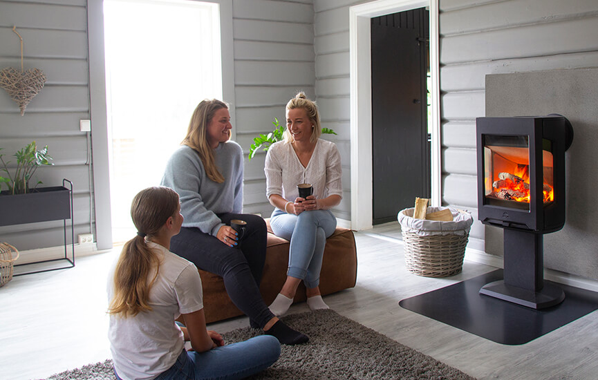 Drei Mädchen sitzen auf einem braunen Hocker in einem grauen Wohnzimmer vor dem Kaminofen Jøtul F 167